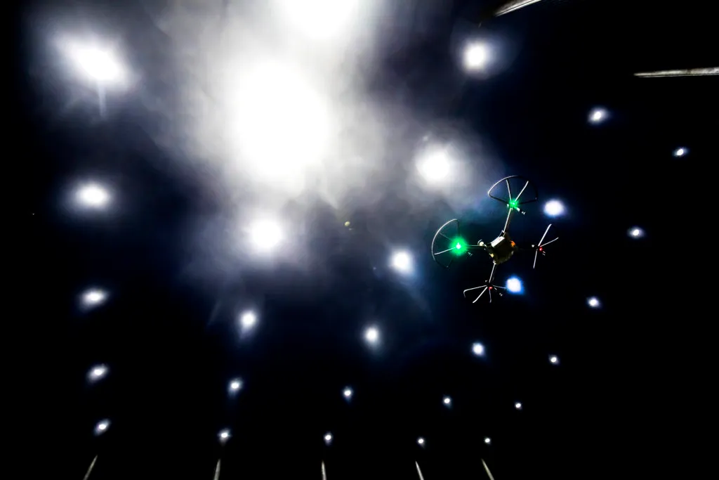 A drone flying at night with navigation lights on under a netted facility with larger spotlights in a grid.