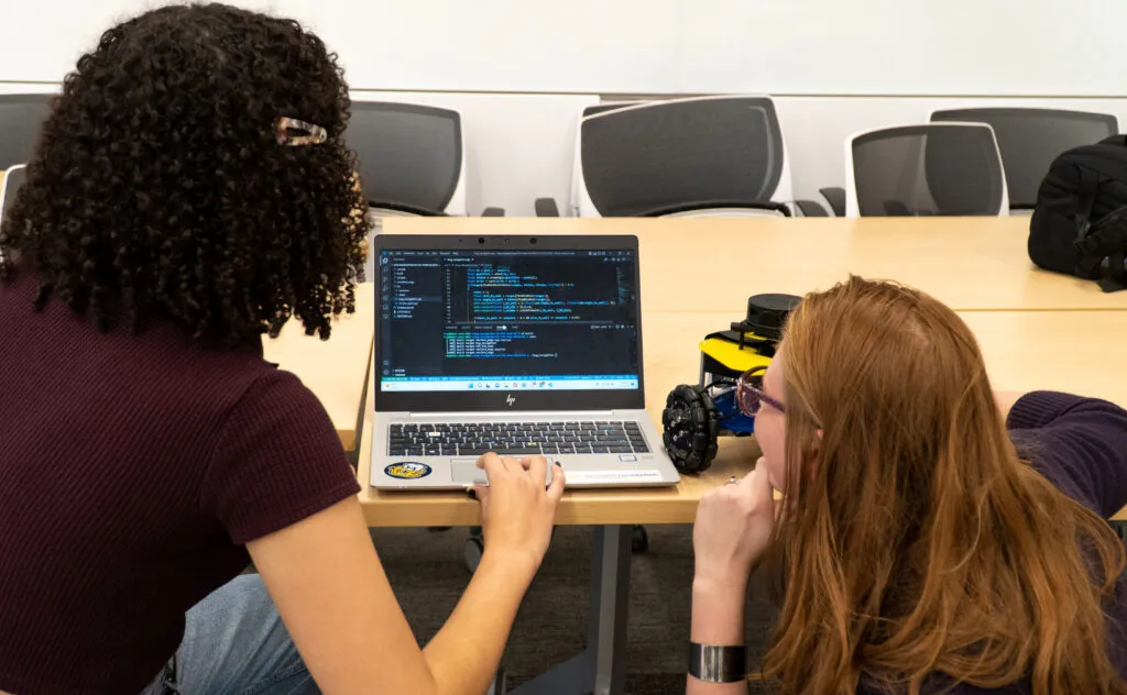 Students in ROB 102 work on coding their Mbot at a table in the classroom.