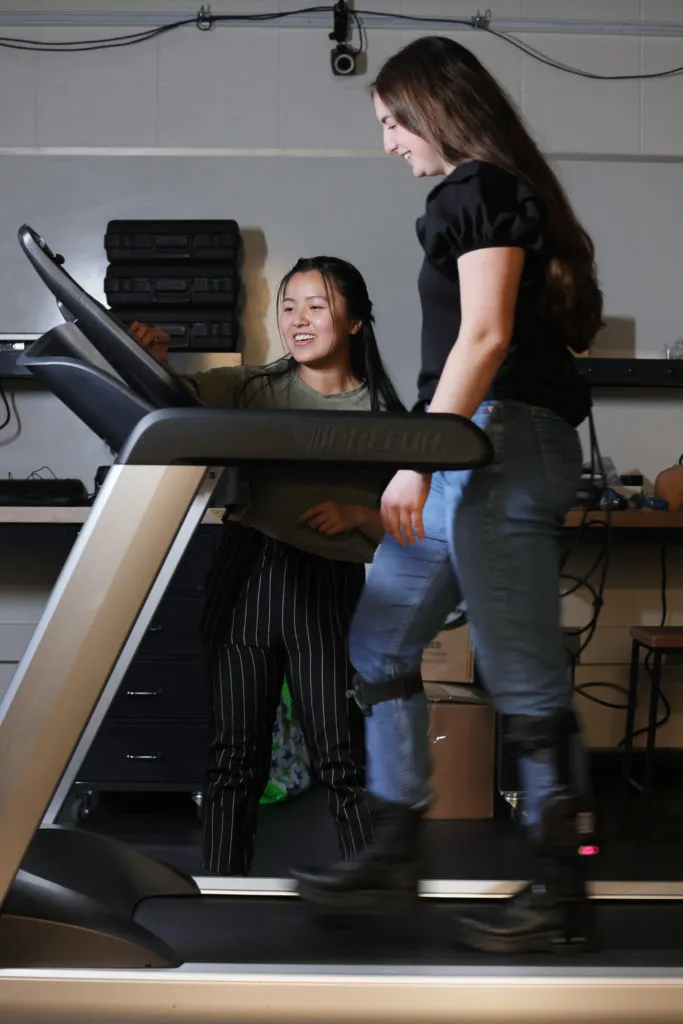 Man I (Maggie) Wu points to a screen on a treadmill as Jacqueline Hannah walks on the treadmill wearing ankle exoskeletons.