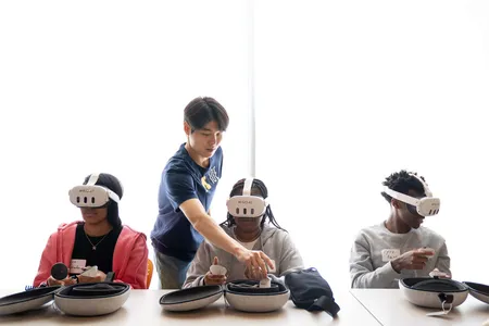 Three high school students sit at a table using VR headsets; one U-M IOE student assists one of the students.