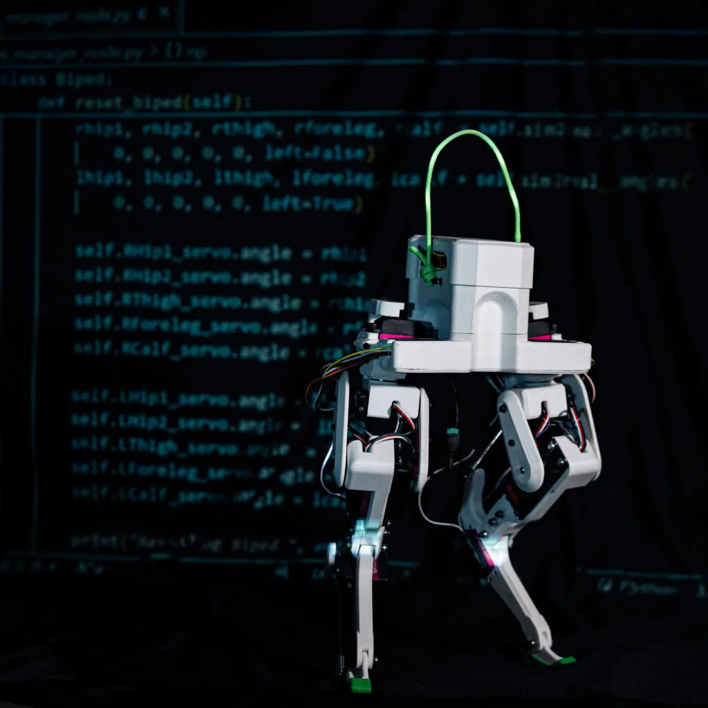 Legolas biped robot stands in front of some of its code on a screen