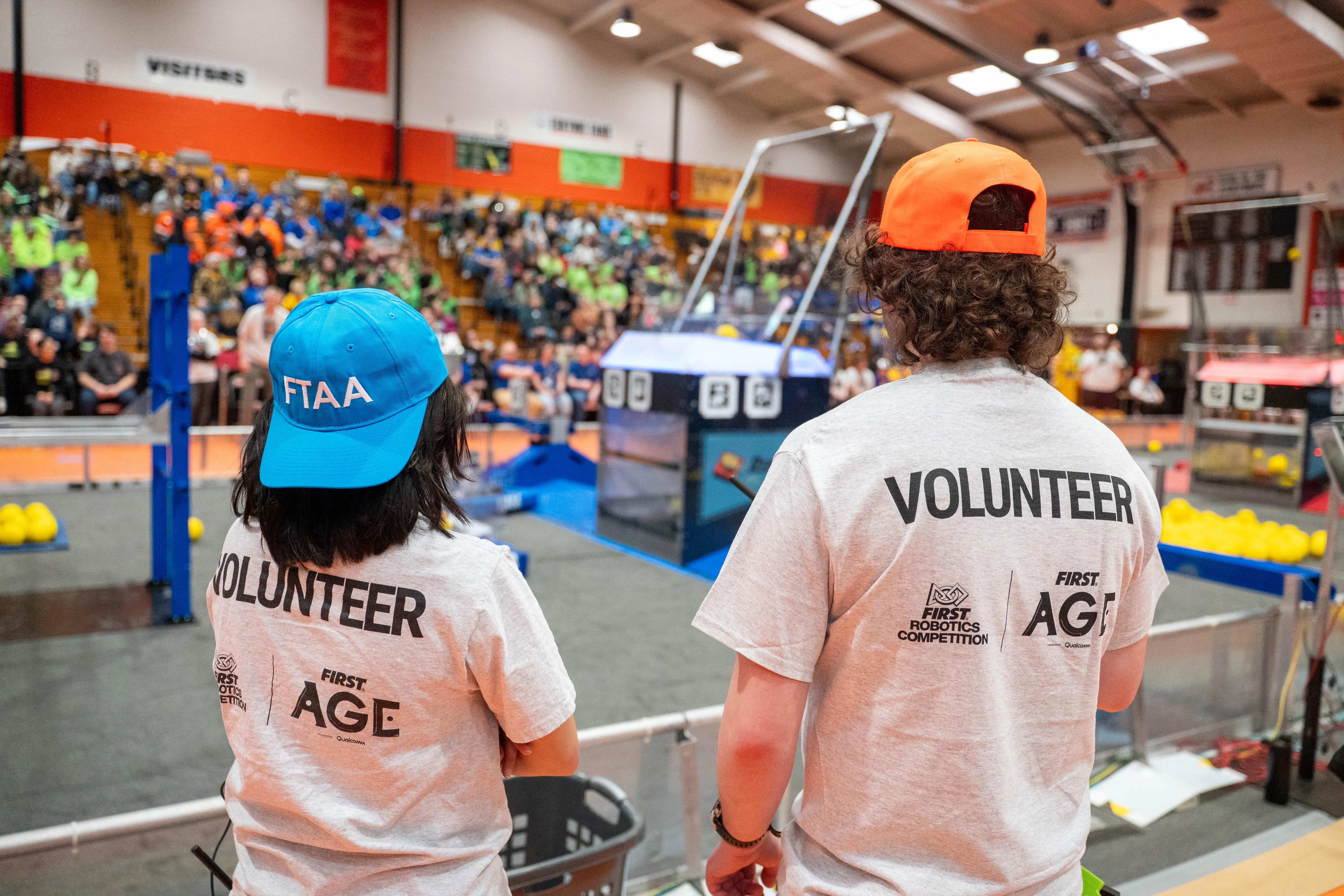 Two volunteers stand with their back toward the camera, one wearing a hat that says FTAA, with a FIRST robotics field in the background and crowd in the stands.