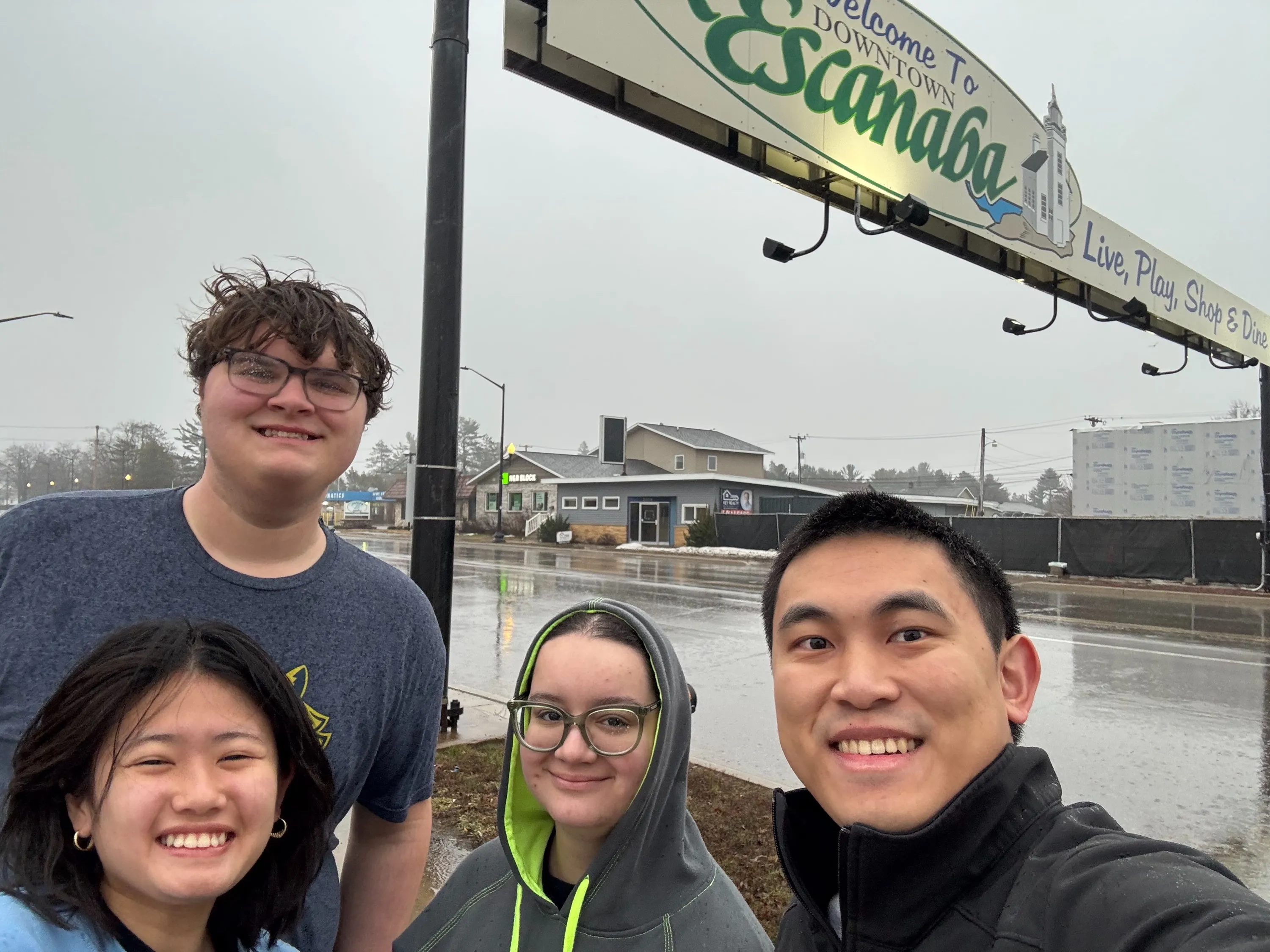 Four FAMNM members standing in rainy weather in front of a sign reading "Welcome to Downtown Escanaba"