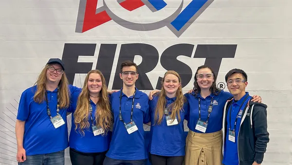 Volunteers pose in front of a FIRST Robotics banner at a competition.