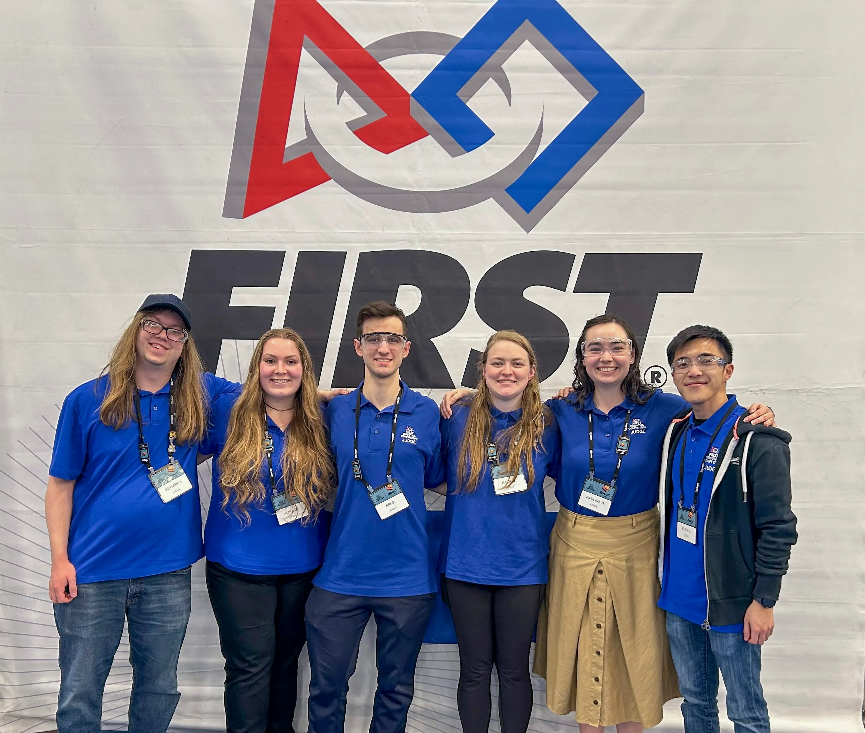 Volunteers pose in front of a FIRST Robotics banner at a competition.