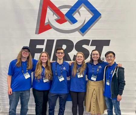 Volunteers pose in front of a FIRST Robotics banner at a competition.