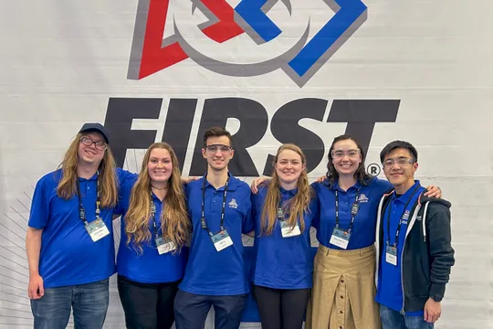 Volunteers pose in front of a FIRST Robotics banner at a competition.