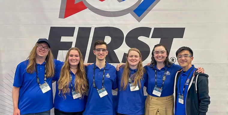 Volunteers pose in front of a FIRST Robotics banner at a competition.