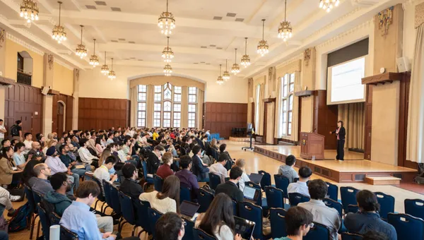 A woman stands on a stage in a ballroom, in front of a large seated audience.