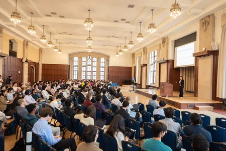 A woman stands on a stage in a ballroom, in front of a large seated audience.