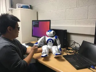 A researcher sits at a desk in front of two computers while extending his hand to the hand of a small humanoid robot, about two feet tall, that sits on the desk.
