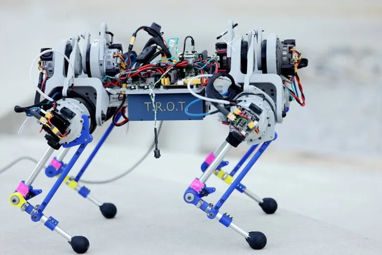 The robot has four legs with backward-facing knees. Each leg has three actuators, controlling hip ab/adduction, hip flexion/extension, and knee flexion/extension.  Exposed wires link up to the motors. T.R.O.T is emblazoned on the case around the belly of the robot.