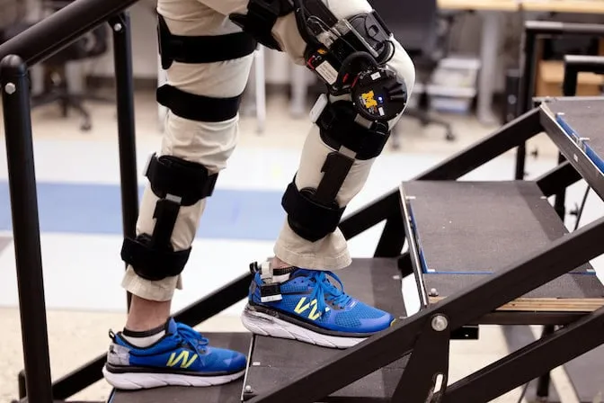 A researcher wearing motorized knee exoskeletons on both legs ascends a flight of stairs.