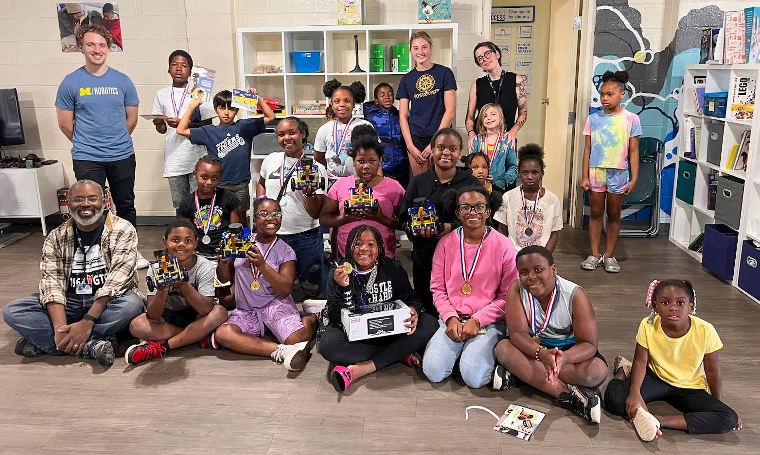 A group of students and instructors pose in a group holding MBot robots up while a few wear golden medallions of completion in a classroom.