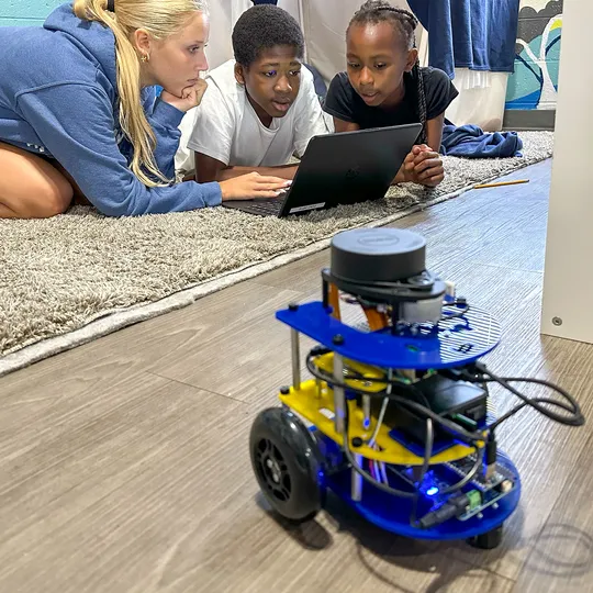 A student works on a laptop with Scratch programming as an MBot robot sets off into a maze made of plastic barriers.