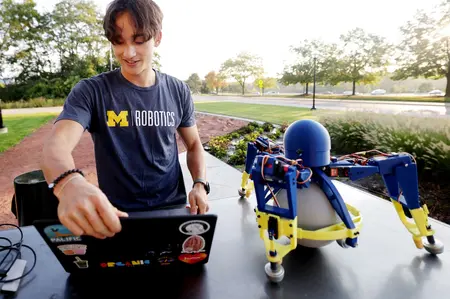A student works on a laptop beside a small legged robot outside the Ford Robotics Building