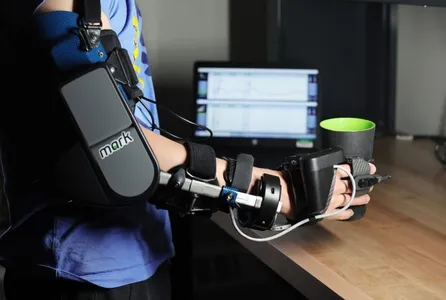 A user with a robotic exoskeleton on their shoulder, forearm, wrist and fingers grasps a cup with a computer logging data in the background.