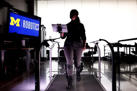 A person walks on a treadmill wearing a robotic exoskeleton in a Michigan Robotics lab