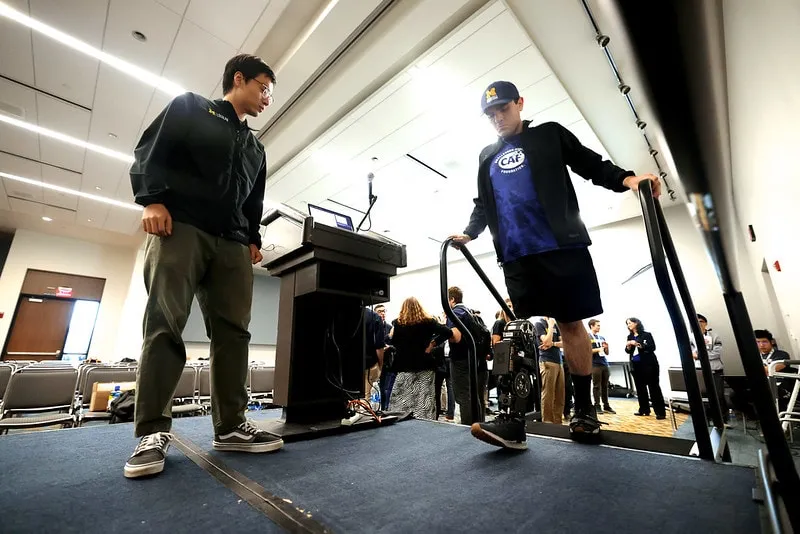 Two people on a platform, one demonstrating use of a prosthetic leg, and the other watching.