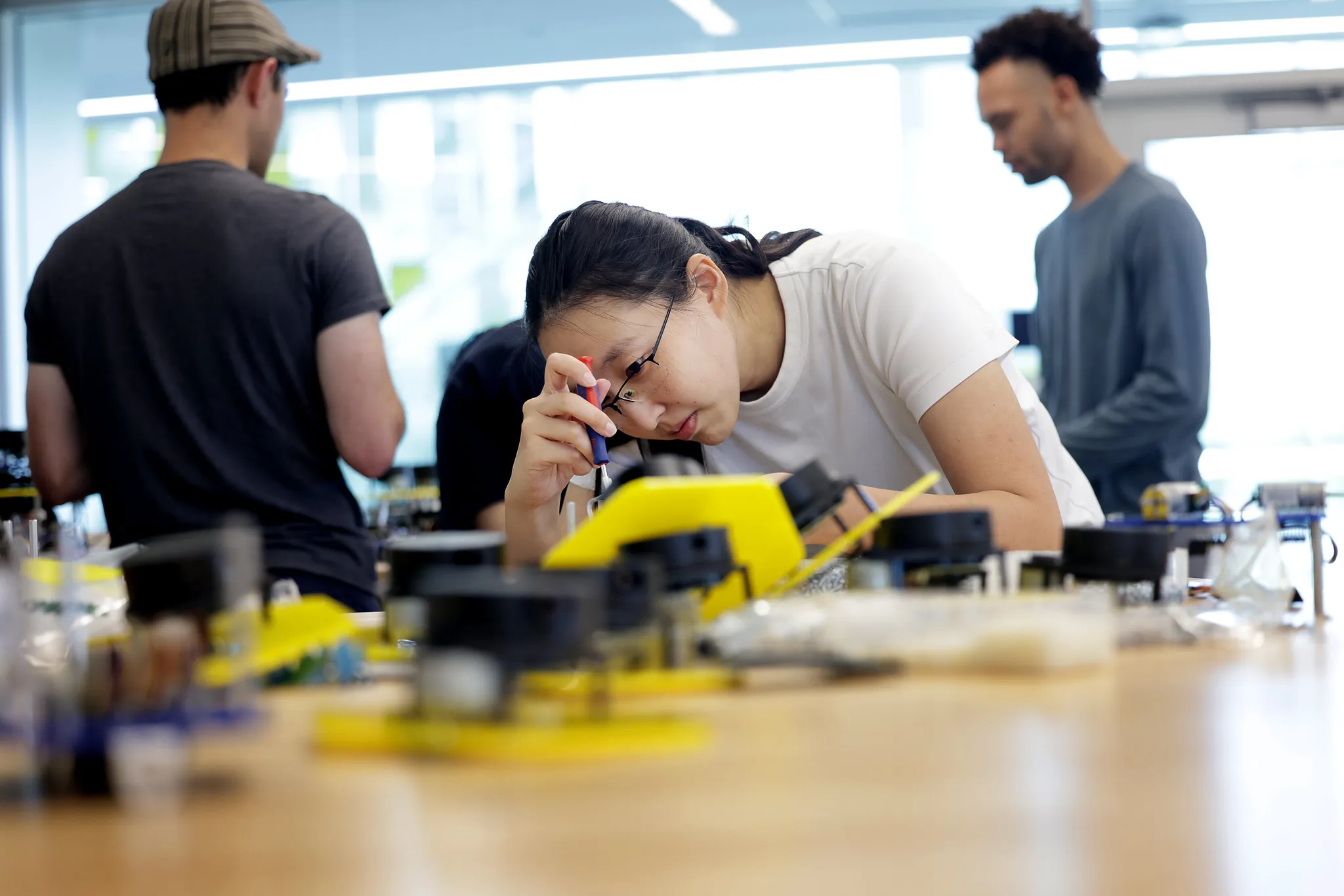 Graduate student instructors assemblying Mbots for upcoming term with 3d printed parts, actuators, and circuit boards scattered around a work table