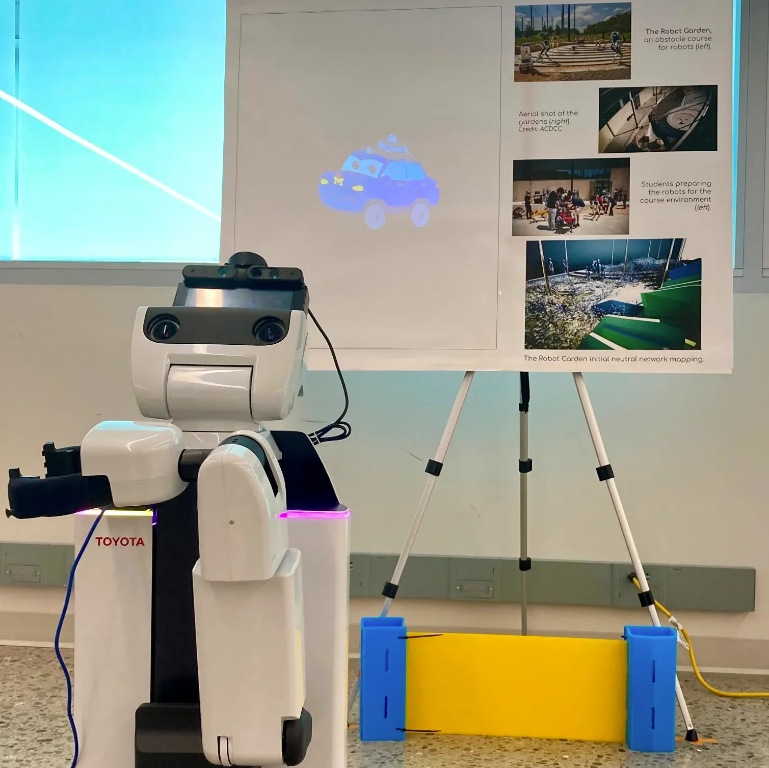 A white Toyota humanoid service robot stands in front of a poster board displaying a projected animated blue car character.