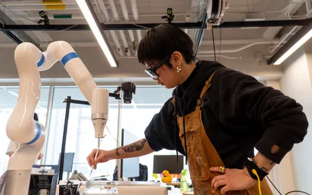 An artist paints opposite of a gleaming white robot arm, also painting on the same canvas in a research lab.