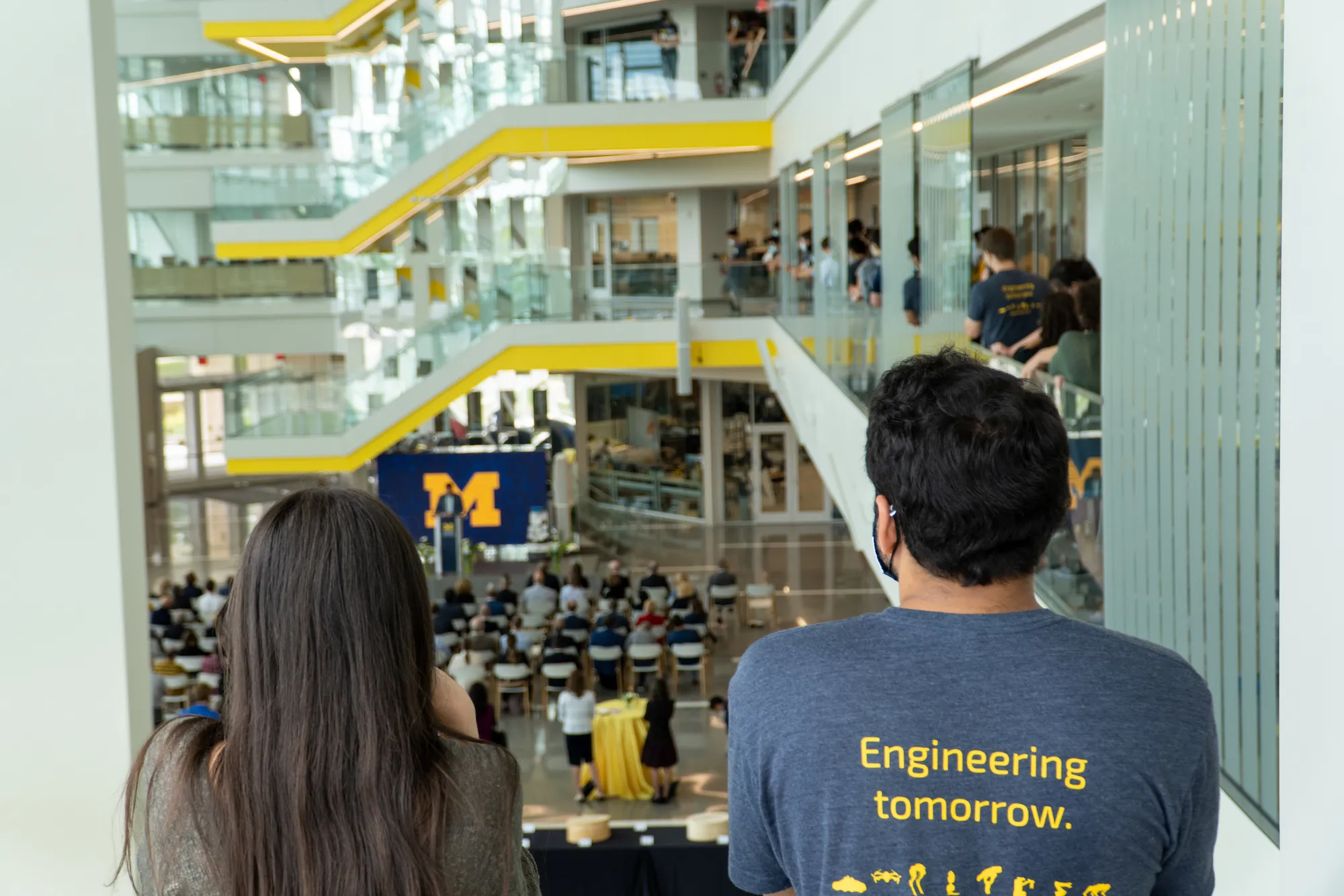 Two students stand looking out over the Robotics Building atrium full of people, one student wearing a shirt that reads 'Engineering Tomorrow' with robot icons on it.