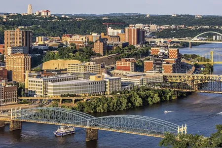 Bridges cross the Monongahela River in Pittsburgh in evening light, with a riverboat passing below and the Cathedral of Learning on the horizon.