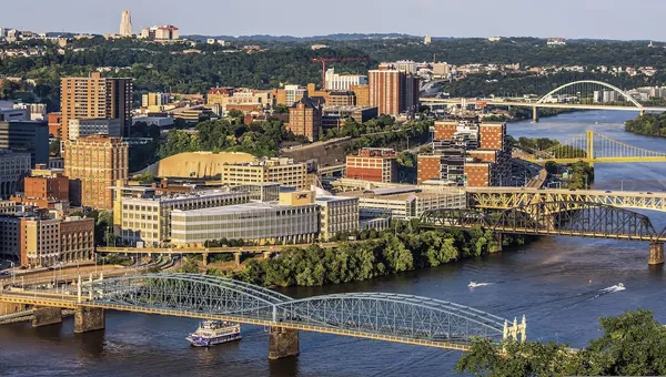 Bridges cross the Monongahela River in Pittsburgh in evening light, with a riverboat passing below and the Cathedral of Learning on the horizon.
