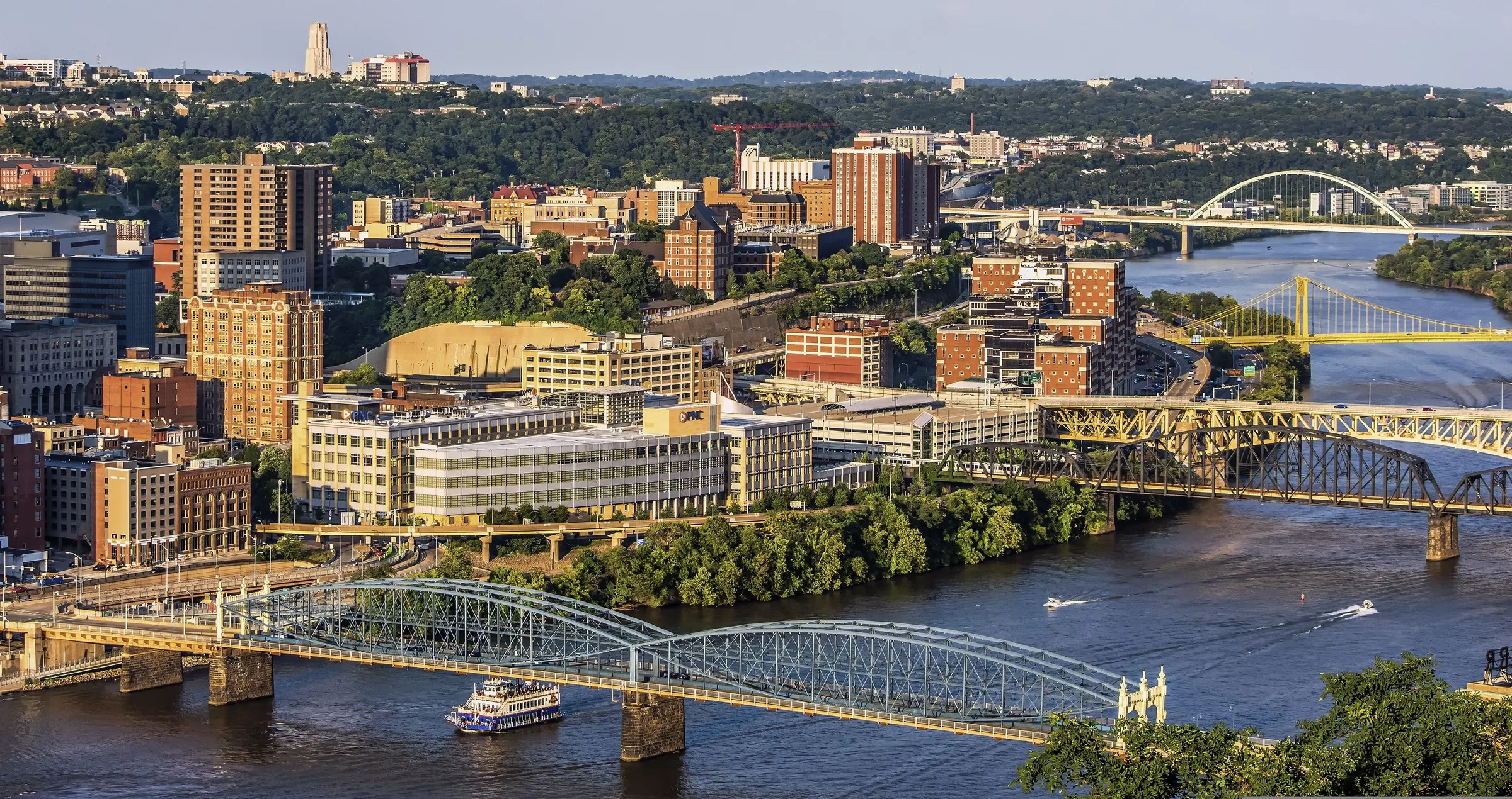 Bridges cross the Monongahela River in Pittsburgh in evening light, with a riverboat passing below and the Cathedral of Learning on the horizon.