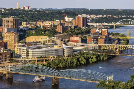 Bridges cross the Monongahela River in Pittsburgh in evening light, with a riverboat passing below and the Cathedral of Learning on the horizon.