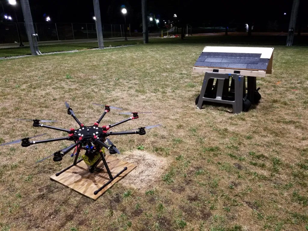 The roofing octocopter, equipped with a nail gun, is parked near the mock roof.