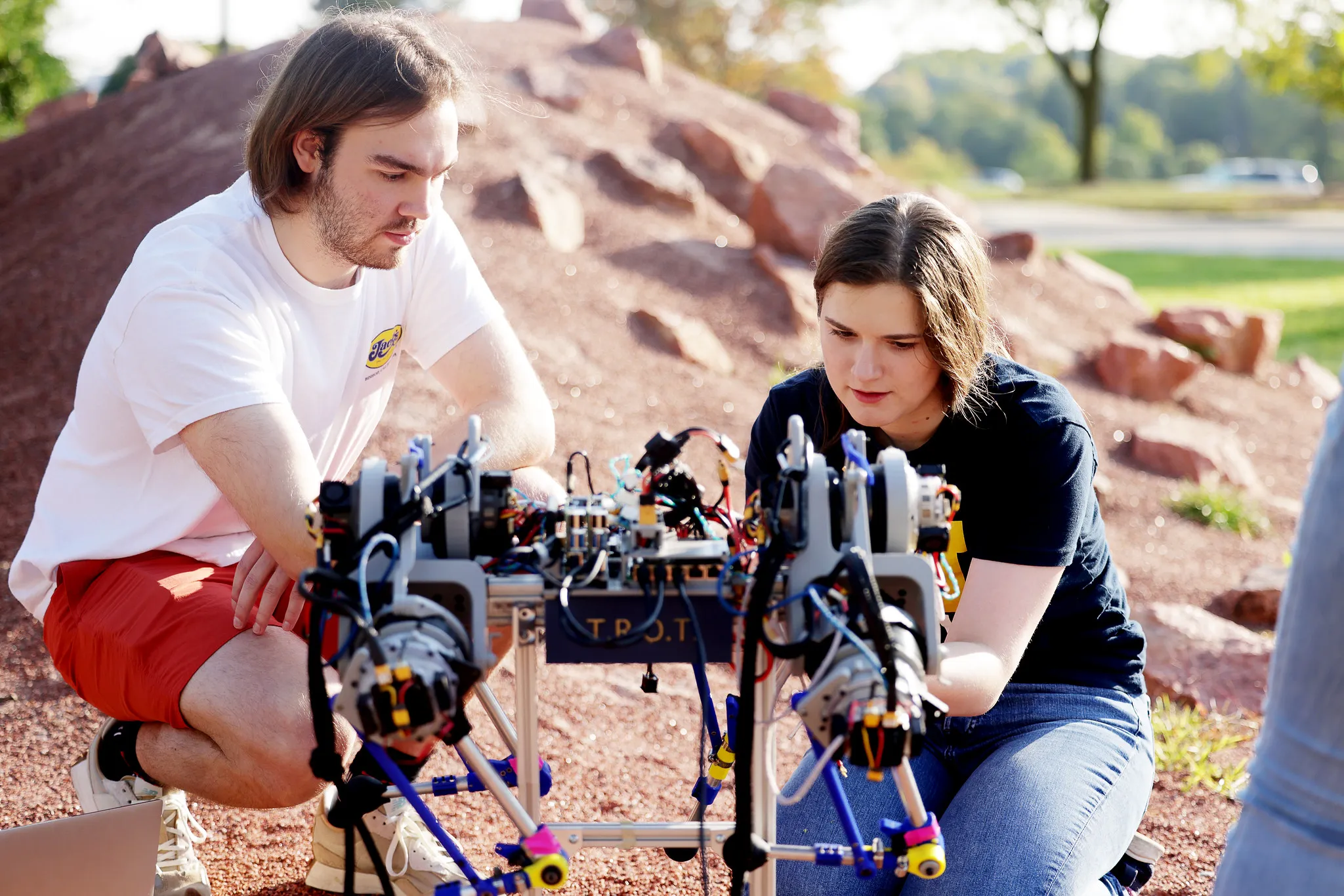 Two graduate students work on a house-built quadruped robot by a large red mound resembling the planet Mars