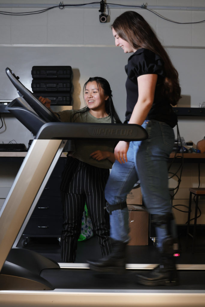 Man I (Maggie) Wu points to a screen on a treadmill as Jacqueline Hannah walks on the treadmill wearing ankle exoskeletons.