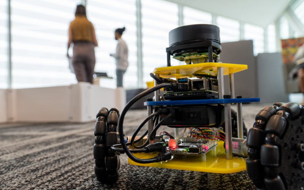 A small robot with three wheels and a LIDAR waits in the foreground with two people in the background.