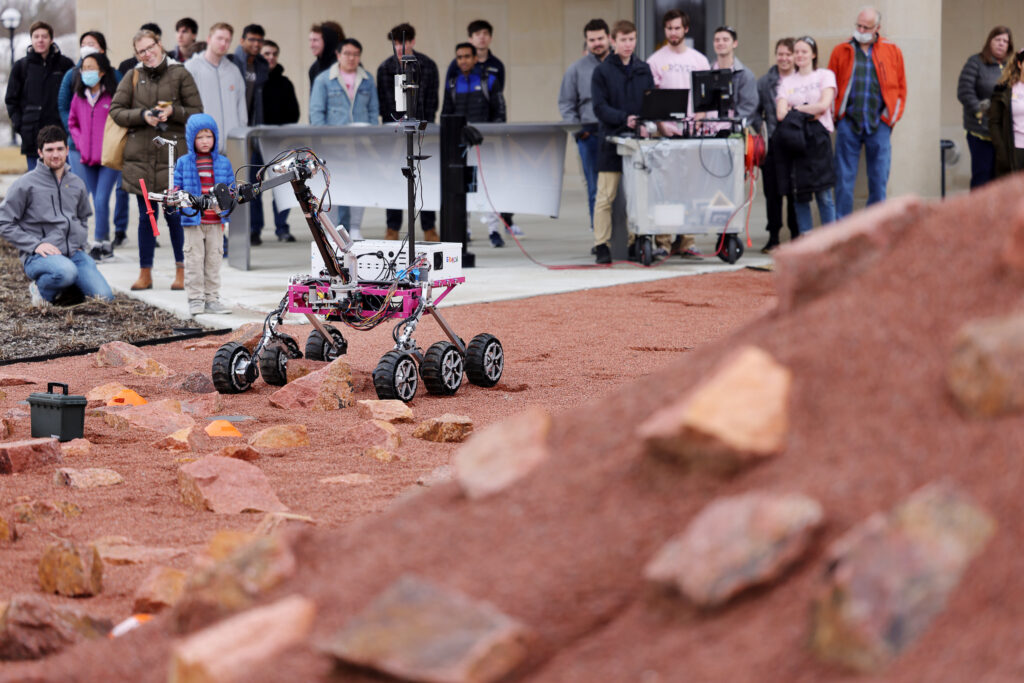 A crowd watches as the Mover robot navigates a Mars-like surface as it is about to grab a box with its robot arm.