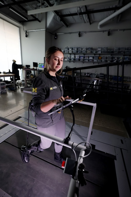 A student stands on a treadmill in a research lab wearing an exoskeleton and selecting power preferences on a tablet.