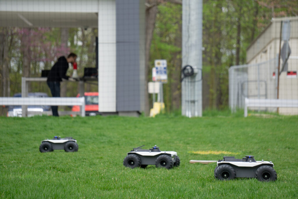 Three four-wheeled robot vehicles drive along grass while a researcher works on a computer in the background in a netted research facility outdoors.