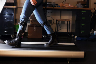 A person wearing sturdy boots and jeans walks on a treadmill wearing a metal brace from the ankle to just below the knee.