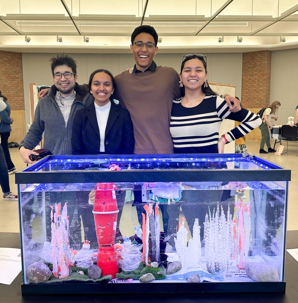 Three students and lecturer stand behind their project of an aquarium filled with robotic fish and plastic pollution decorated to be an aquatic environment.