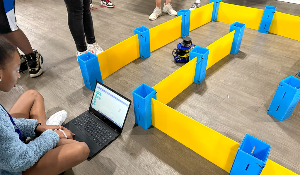 A student works on a laptop with Scratch programming as an MBot robot sets off into a maze made of plastic barriers.