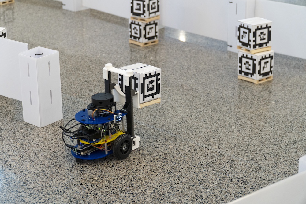 An MBot educational robot with two wheels and a forklift carries a small box on a pallet covered in April Tags, QR-Code like figures, as it goes to stack it on other boxes.